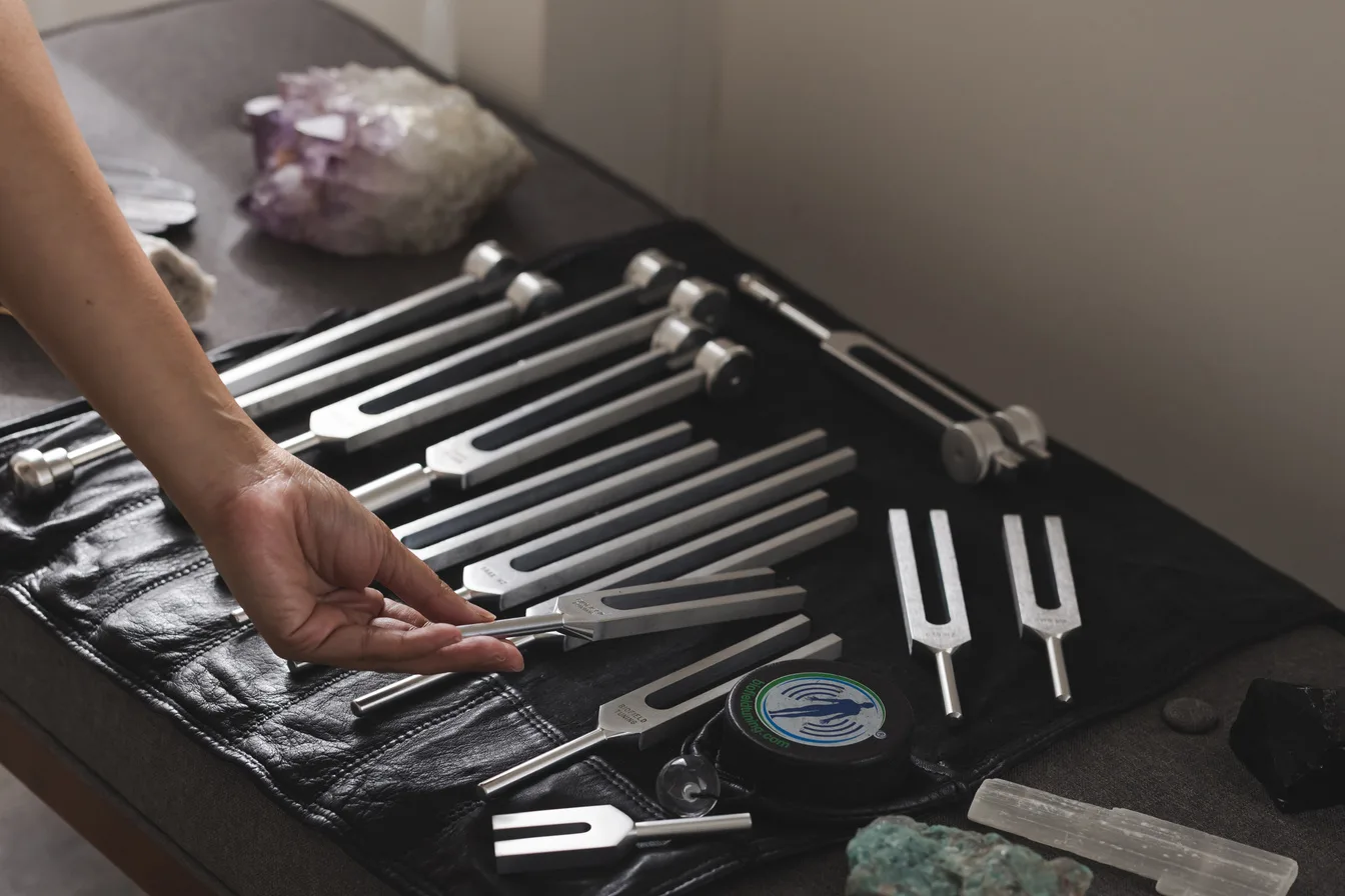 Vanessa's hand selecting a calibrated tuning fork from a full set laid out alongside crystals