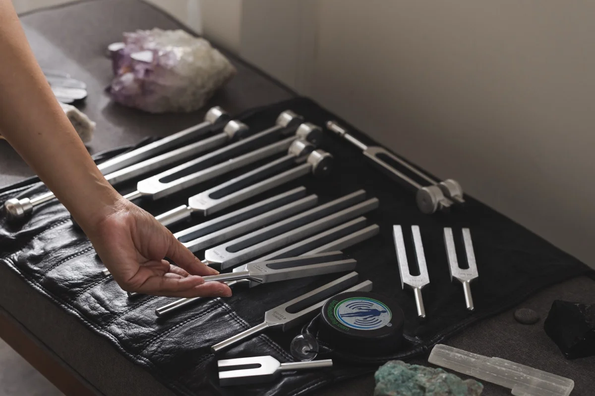 Vanessa's hand selecting a calibrated tuning fork from a full set laid out alongside crystals